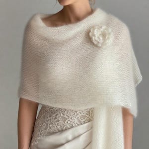 May include: Ivory-colored, knitted shawl with a flower accent. The shawl is draped over a dress, creating a soft, textured look. The flower is a matching ivory color, adding a delicate touch to the bridal or formal wear.