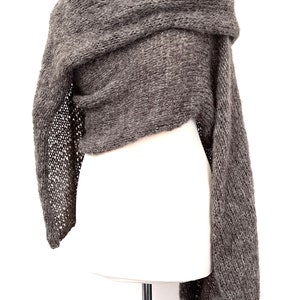 May include: A gray knitted shawl draped over a white mannequin. The shawl has a textured appearance with a loose, open-knit design on the edges. The fabric appears soft and warm, suitable for cooler weather.