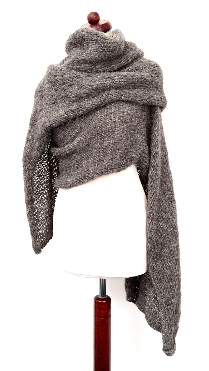 May include: A gray knitted shawl draped over a white mannequin. The shawl has a textured appearance with a loose, open-knit design on the edges. The fabric appears soft and warm, suitable for cooler weather.