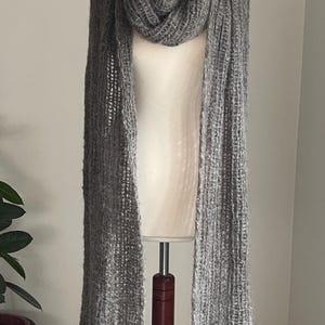 May include: A long, gray scarf draped over a mannequin. The scarf is made of a thick, textured knit material. The scarf is displayed on a wooden stand. The background is a plain, light-colored wall.