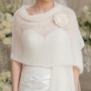 May include: A white, sheer, knitted shawl with a flower accent. The shawl is draped over a white wedding dress. The flower is a matching off-white color. The bride is wearing a veil.