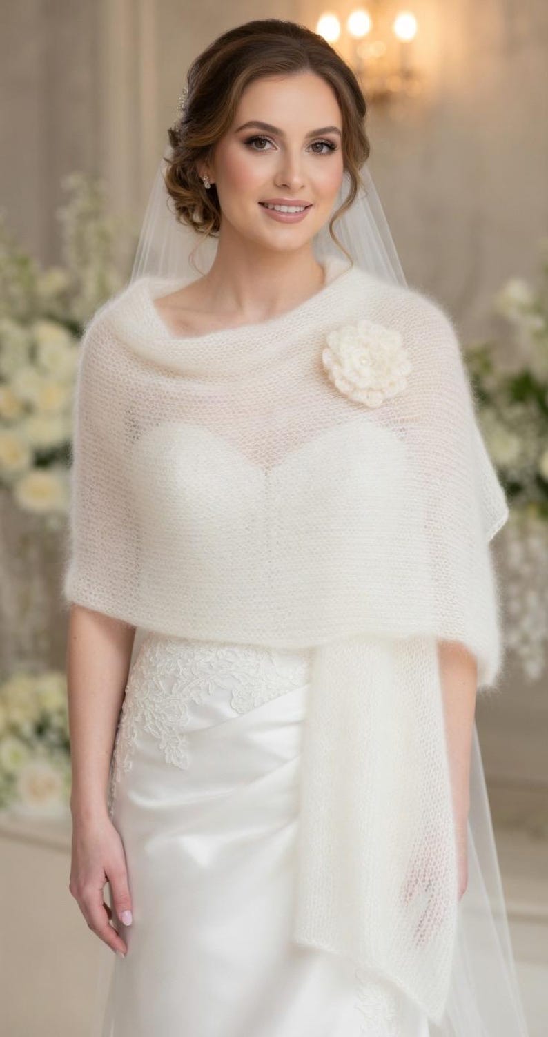 May include: A white, sheer, knitted shawl with a flower accent. The shawl is draped over a white wedding dress. The flower is a matching off-white color. The bride is wearing a veil.