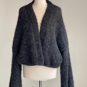 May include: A charcoal grey, open-front cardigan with long, voluminous sleeves. The sweater is displayed on a mannequin, highlighting its soft, textured knit. The V-neck design and relaxed fit create a cosy, stylish look.
