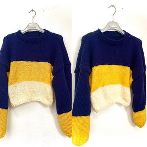 May include: A colour-blocked cropped sweater in navy blue, yellow, and off-white. The top and cuffs are navy, with a yellow stripe in the middle and an off-white stripe at the bottom. The sweater is hanging on a silver hanger.