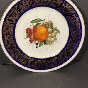 May include: A white ceramic plate with a blue and gold rim. The center of the plate features a painted image of fruit, including an orange, grapes, and raspberries.