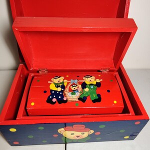May include: A red wooden toy chest with a blue and white polka dot design. The lid is open and reveals a smaller red chest with a painted scene of three bears wearing hats and holding a basket. The smaller chest has a yellow lid with red polka dots.