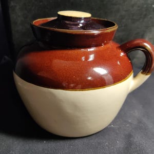 May include: A brown and white ceramic crock with a lid. The crock has a brown band around the middle and a brown handle.