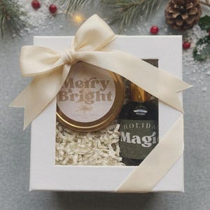 May include: A white square gift box with a clear window, tied with a cream-colored ribbon. Inside, a gold-lidded candle labeled Merry & Bright and a dark glass bottle labeled Holiday Magic rest on a bed of shredded white paper filler.