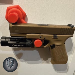 Könnte beinhalten: Eine braune Pistole mit einer schwarzen Taschenlampe, die an der Unterseite befestigt ist. Die Waffe ist an einer roten Wandhalterung aus Kunststoff befestigt. Die Waffe trägt die Aufschrift "GLOCK 19X" und "AUSTRIA" an der Seite.