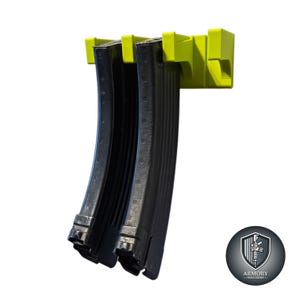 Puede incluir: Un soporte de pared para cargadores de color verde brillante con dos cargadores negros unidos. El soporte tiene un logotipo que dice "Armory Solutions".