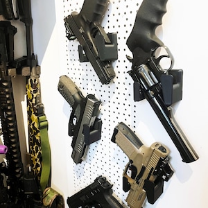 Puede incluir: Una colección de pistolas montadas en una pared perforada blanca. Las armas de fuego son de varios tamaños y colores, incluyendo negro, gris y beige. La imagen muestra una variedad de diseños y estilos de pistolas.