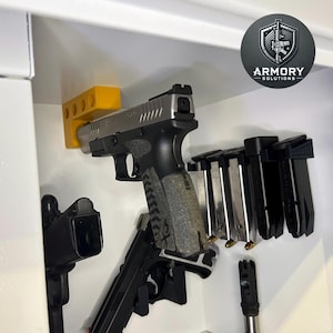 May include: A display of firearms and magazines inside a white cabinet. The image features a silver and black handgun, several magazines, and a yellow holder. The Armory Solutions logo is visible in the upper right corner.