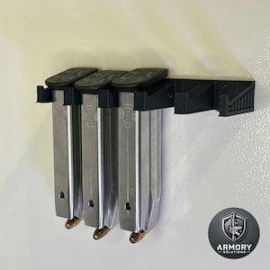 Puede incluir: Un soporte para cargadores de plástico negro con tres cargadores plateados y un logotipo en blanco y negro que dice "Armory Solutions".