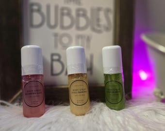 May include: Three small body spray bottles with white caps. The bottles contain pink, yellow, and green liquids. Each bottle has a label with the words "Body Spray" and a flavor name. A sign in the background reads "You are the bubbles to my bath."