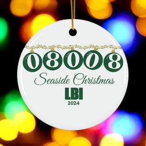 May include: A white ceramic Christmas ornament with a gold string. The ornament features the numbers "08008" in green, the words "Seaside Christmas", "LBI", and the year "2024". A green and red garland decorates the top.