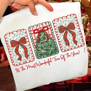 Puede incluir: Bolso tote blanco con temática navideña con tres diseños rectangulares en rojo y verde. Los diseños incluyen un árbol de Navidad y lazos a cuadros. El texto "It's The Most Wonderful Time Of The Year!" está impreso en rojo.