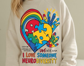 May include: A beige sweatshirt with a rainbow and a heart made of puzzle pieces. The text on the shirt reads "Everyone communicates differently. I love with autism. I love someone neurodiversity."