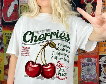 May include: White t-shirt with a graphic of two red cherries and the text "Cherries" in green. The text "Fruit of the Spirit" is above the cherries. Below the cherries are the words "Kindness, Goodness, Faithfulness, Self-control, Love, Joy, Peace, Forbearance".