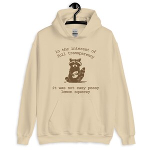Funny graphic hoodie featuring a sketched raccoon and sarcastic quote about life not being easy. Beige oversized sweatshirt with dry humor for mental overload and adulting exhaustion. Great unisex hoodie for students, burned-out professionals, or anyone who loves ironic quotes. Cozy fashion with a relatable twist, perfect for everyday streetwear or as a humorous gift.