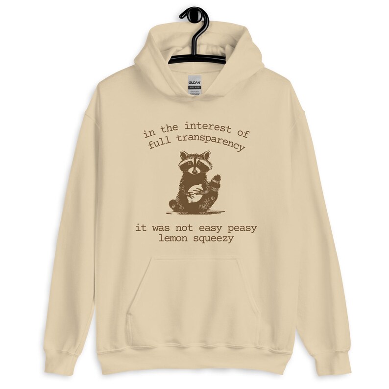 Funny graphic hoodie featuring a sketched raccoon and sarcastic quote about life not being easy. Beige oversized sweatshirt with dry humor for mental overload and adulting exhaustion. Great unisex hoodie for students, burned-out professionals, or anyone who loves ironic quotes. Cozy fashion with a relatable twist, perfect for everyday streetwear or as a humorous gift.