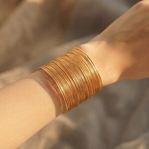 May include: A wide cuff bracelet composed of numerous thin, stacked gold-toned wire bangles. The piece features a continuous, uniform metallic texture that wraps around the wrist, creating a layered appearance with a polished, reflective finish.
