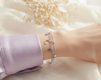 May include: A multi-strand silver bracelet featuring delicate chain links, small metallic beads, and dangling charms in the shapes of a crescent moon and stars. The bracelet is worn on a wrist, showcasing the polished silver finish and the layered design.