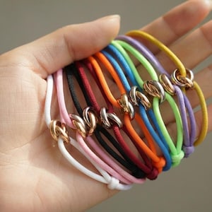 Könnte beinhalten: Eine Sammlung von elastischen Haargummis in verschiedenen Farben, darunter Weiß, Rosa, Schwarz, Rot, Orange, Blau, Grün, Lila und Gelb. Jedes Haargummi verfügt über einen metallischen, knotenförmigen Anhänger in polierter Goldoptik.