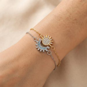 May include: A two-strand bracelet featuring a gold-toned sun charm encrusted with clear crystals and a silver-toned crescent moon charm adorned with light blue and clear crystals. The delicate metal chains connect to each celestial-themed pendant.