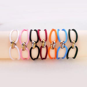 May include: Seven hair ties in white, pink, black, maroon, orange, blue, and dark green. Each elastic band features a central decorative element consisting of three interlocking metal rings in gold, silver, and rose gold tones.