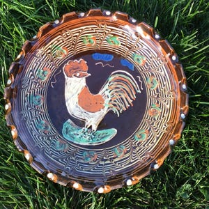 May include: A decorative ceramic plate with a rooster design. The plate features a dark brown center with a white and orange rooster illustration. The rim is brown with white dots and a patterned design. The plate is on a bed of green grass.