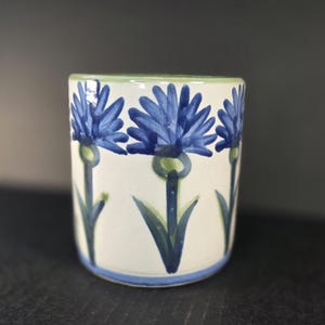 May include: A cylindrical ceramic mug with a cream-colored base featuring a repeating hand-painted pattern of blue cornflowers with green stems and leaves. The rim and base are accented with thin blue and green horizontal painted bands.
