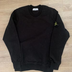 Könnte beinhalten: Ein schwarzes Langarm-Sweatshirt mit Rundhalsausschnitt sowie gerippten Bündchen an Ärmeln und Saum. Das Kleidungsstück verfügt über ein weißes Innenetikett mit der Aufschrift Stone Island und ein Kompass-Logo am linken Oberarm.