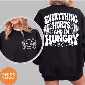 May include: A black sweatshirt featuring a white graphic on the back that reads "EVERYTHING HURTS AND I'M HUNGRY" with a barbell and cutlery. The front chest area displays a white graphic of a smiling emoji holding a fork and a knife.