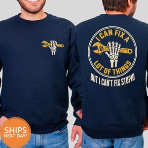 May include: A navy blue crewneck sweatshirt featuring a small graphic on the front left chest of a skeleton hand holding a yellow wrench. The back displays a large circular graphic with a skeleton hand holding a wrench and the text I CAN FIX A LOT OF THINGS BUT I CAN'T FIX STUPID.