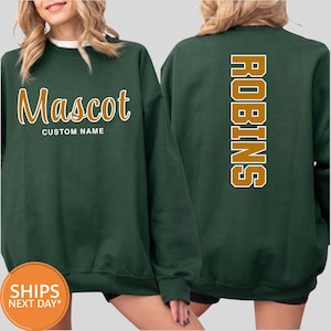 May include: A dark green crewneck sweatshirt featuring the word Mascot in a gold script font on the front, with the text CUSTOM NAME below it. The back of the sweatshirt displays the word ROBINS in large, vertical gold block letters.