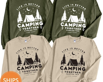 Pode incluir: Quatro camisolas de gola redonda em verde azeitona e bege com uma impressão gráfica de uma tenda, fogueira, árvores e uma lua crescente. O texto diz: Life is better camping together, 2026 camping trip, Lake Tahoe. A impressão é branca.