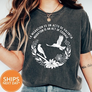May include: Dark gray t-shirt with white graphic of two birds inside a floral wreath, with the text "LA MIGRACION ES UN ACTO DE VALENTIA MIGRATION IS AN ACT OF COURAGE". The shirt is a comfortable, casual style.