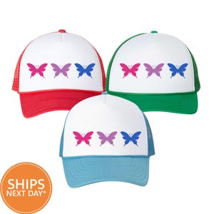 May include: Three trucker hats with white front panels and mesh backs. Each hat features a row of three butterflies in pink, purple, and blue. The hats have red, green, and light blue brims and mesh respectively, with a structured front panel.