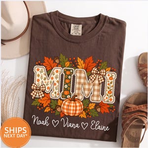 May include: A brown short-sleeve t-shirt featuring the text MIMI in large letters decorated with floral and plaid patterns. The design includes autumn leaves, acorns, and pumpkins. Below the graphic, the names Noah, Viana, and Elaine are printed.
