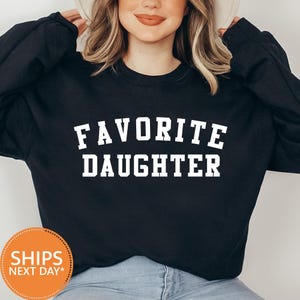 May include: Black sweatshirt with the text "FAVORITE DAUGHTER" in white block letters. The sweatshirt has long sleeves and a crew neck. The image also includes a round orange sticker with the text "SHIPS NEXT DAY*".