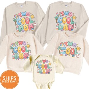 May include: A collection of beige long-sleeved sweatshirts and a baby bodysuit featuring colorful summer-themed graphics. Each garment displays the text "TWO COOL" followed by "MOM," "DAD," "BRO," or "SIS," surrounded by palm trees, suns, and beach items.