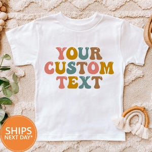 May include: White t-shirt with the text "YOUR CUSTOM TEXT" in a retro font. The text is in various colors, including brown, yellow, pink, teal, and orange. The shirt is a children's size.