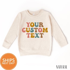May include: A cream-colored sweatshirt with the text "YOUR CUSTOM TEXT" in colorful, retro-style lettering. The sweatshirt has long sleeves and a crew neck. The word "NATURAL" is printed in black at the bottom right.