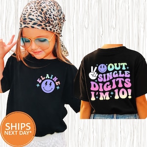 May include: Black t-shirts with colorful graphics. One shirt has a smiley face with the name "Elaine" and lightning bolts. The other shirt says "Out Single Digits I'm 10!" with a peace sign and smiley face.