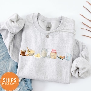 May include: A light gray crewneck sweatshirt featuring a graphic illustration of baking ingredients including a brown sugar bag, a bowl with a wooden spoon, butter cubes, a glass jar of flour, a flour bag, eggs, and a measuring cup.
