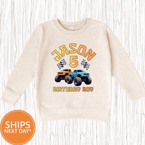 May include: A beige sweatshirt with a graphic featuring the name "Jason", the number "5", and two monster trucks. The text "BIRTHDAY BOY" is below the trucks. Checkered flags are on either side of the number 5.