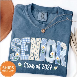 May include: A blue, pigment-dyed t-shirt featuring the word SENIOR in large, block-style letters filled with various blue and white patterns, including gingham, stripes, and daisy prints. Below, the text Class of 2027 is printed in white.