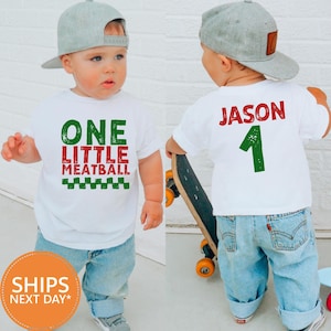 May include: A white t-shirt with the text "ONE LITTLE MEATBALL" in green and red, and a green and white checkered design. The back of the shirt has the name "JASON" and the number "1" in green. The child is wearing a gray baseball cap and blue jeans.