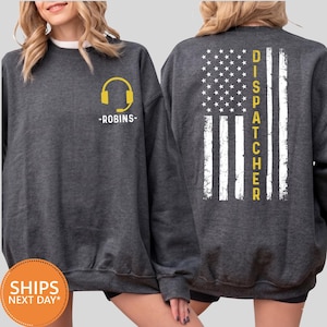 May include: A dark charcoal gray crewneck sweatshirt featuring a yellow headset icon and the text ROBINS on the front left chest. The back displays a distressed white American flag graphic with the word DISPATCHER printed vertically in yellow.