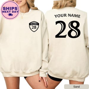 May include: A sand-colored sweatshirt with a black shield-shaped patch on the left chest that says "NAME 28". The back of the sweatshirt has the text "YOUR NAME" and the number "28" in a large, bold font.